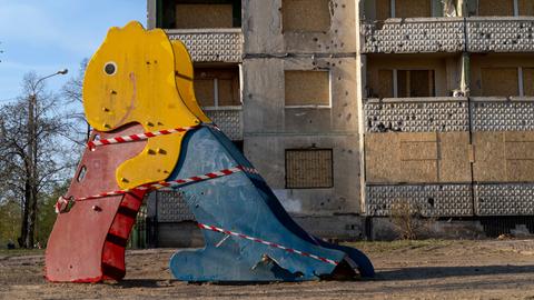 Auf einem Spielplatz steht eine beschädigte bunte Rutsche in Form eines Dinosauriers, im Hintergrund ist ein vom Krieg stark beschädigtes Gebäude zu sehen.