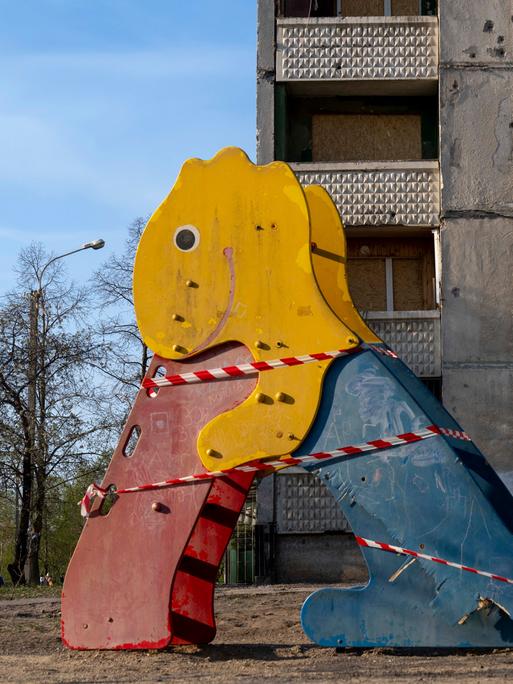 Auf einem Spielplatz steht eine beschädigte bunte Rutsche in Form eines Dinosauriers, im Hintergrund ist ein vom Krieg stark beschädigtes Gebäude zu sehen.