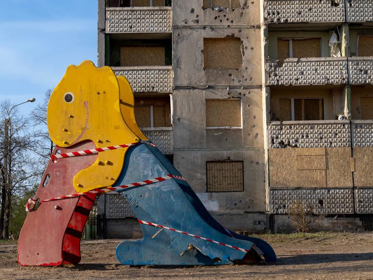 Auf einem Spielplatz steht eine beschädigte bunte Rutsche in Form eines Dinosauriers, im Hintergrund ist ein vom Krieg stark beschädigtes Gebäude zu sehen.