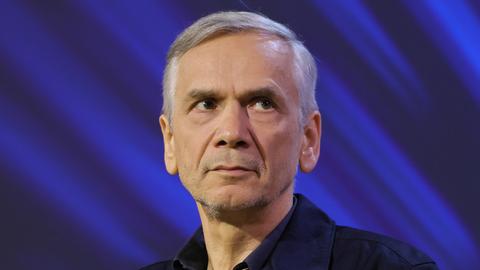 Lutz Seiler sitzt auf einer Bühne mit blauem Hintergrund und schaut leicht zur Seite.