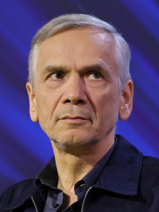 Lutz Seiler sitzt auf einer Bühne mit blauem Hintergrund und schaut leicht zur Seite.