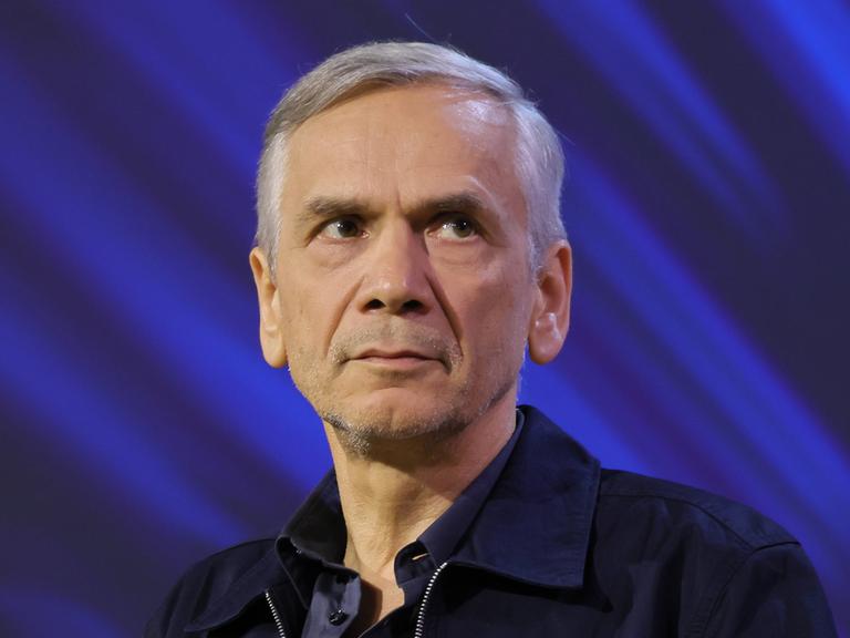 Lutz Seiler sitzt auf einer Bühne mit blauem Hintergrund und schaut leicht zur Seite.