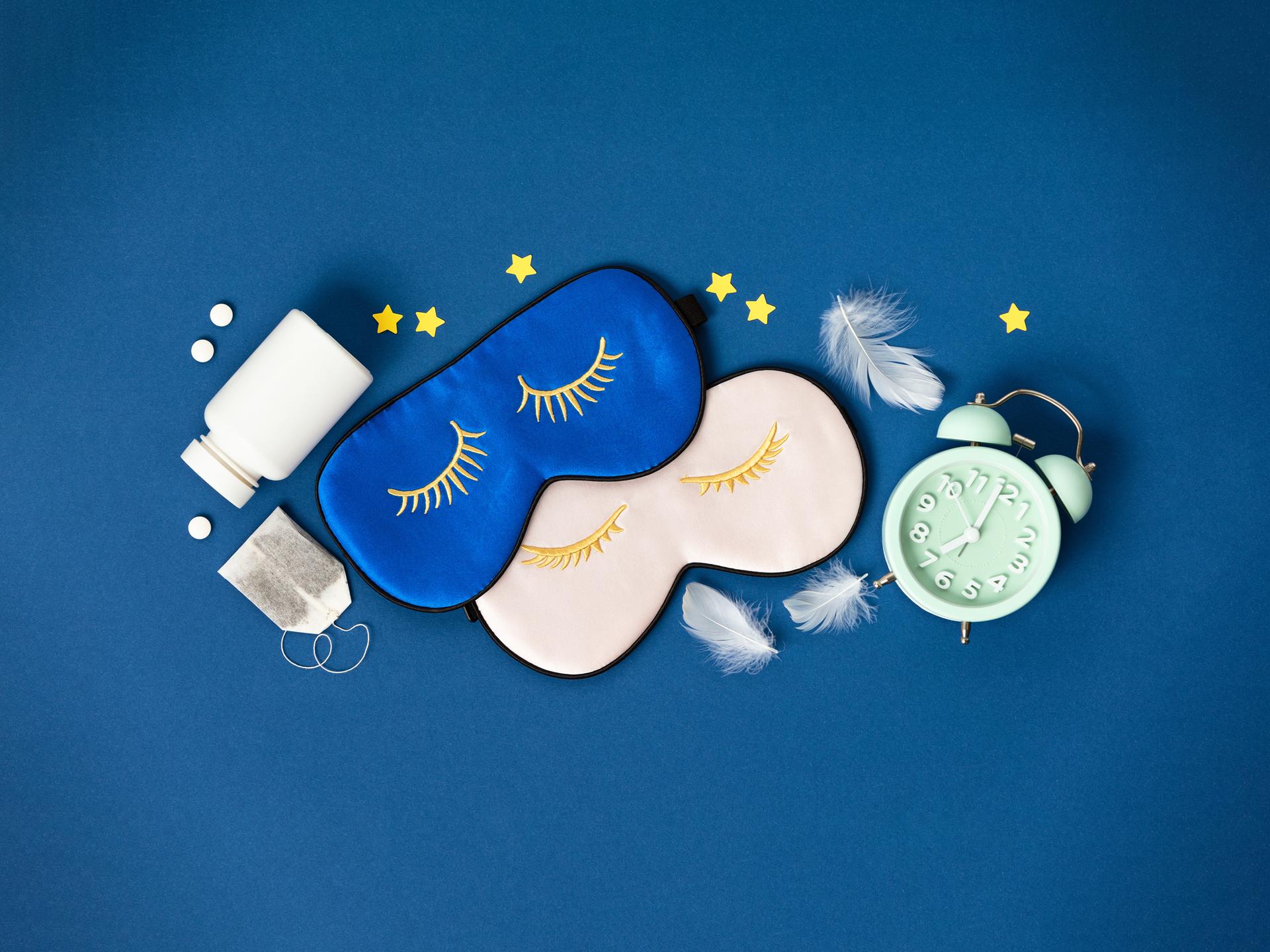 Blaue und rosa Schlafmasken, Tabletten und Kräutertee liegen auf blauem Hintergrund.