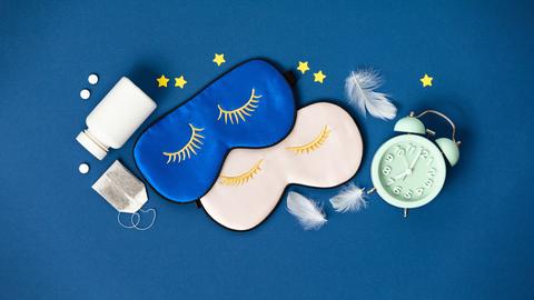 Blaue und rosa Schlafmasken, Tabletten und Kräutertee liegen auf blauem Hintergrund.