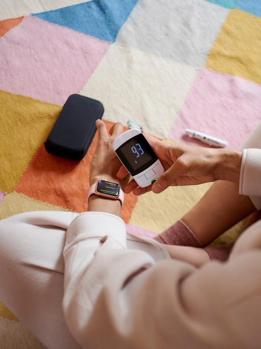 Eine junge Frau überprüft ihren Blutzucker mit einem Messgerät und versucht, ihn mit einer Smartwatch-App zu synchronisieren.