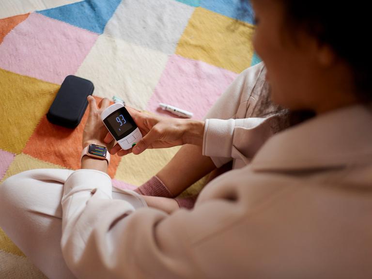 Eine junge Frau überprüft ihren Blutzucker mit einem Messgerät und versucht, ihn mit einer Smartwatch-App zu synchronisieren.