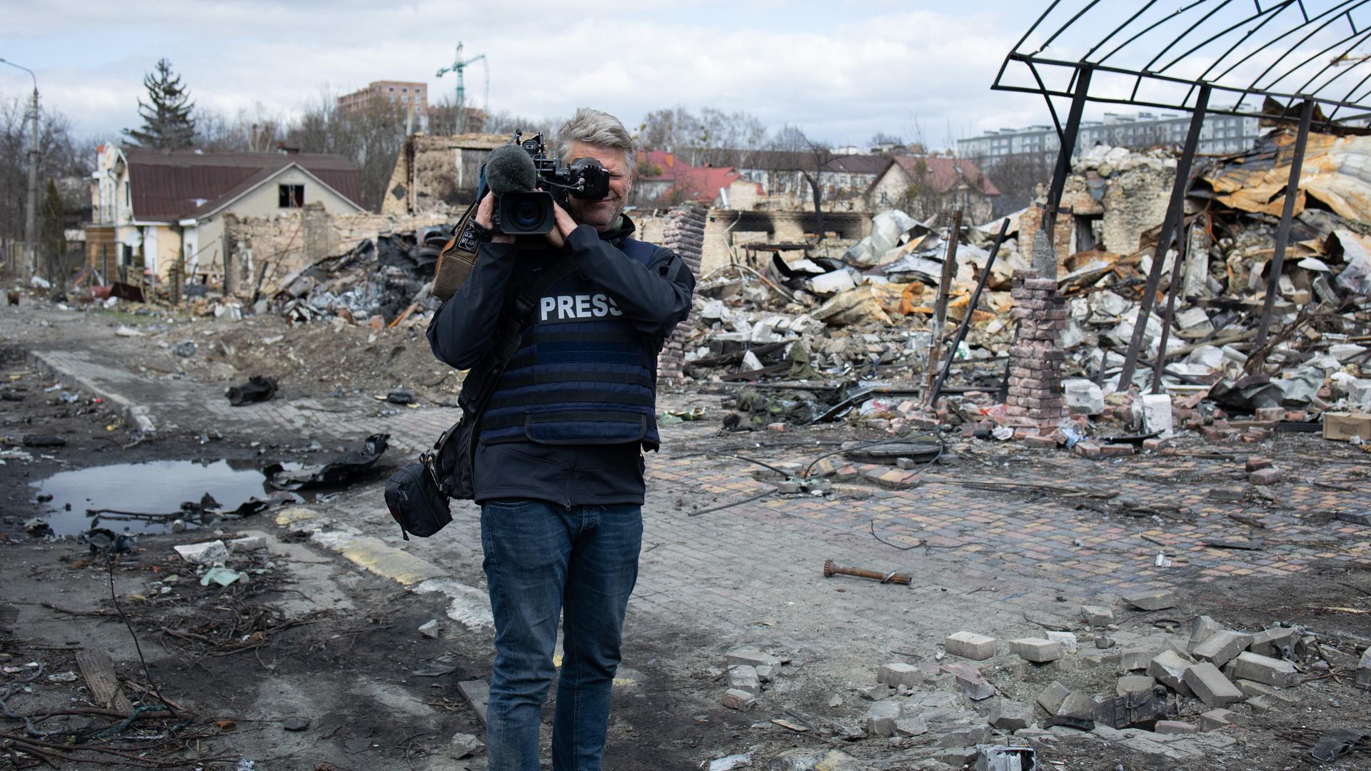Das Foto zeigt einen Reporter mit Kamera auf der Schulter, dahinter sieht man zerstörte Gebäude.