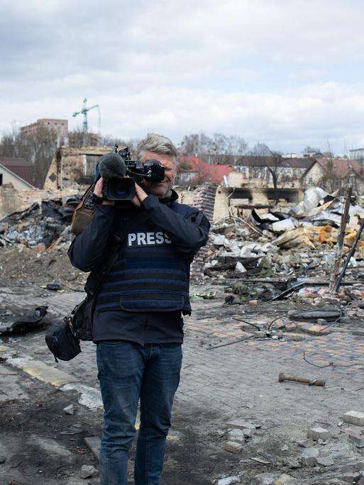 Ein Journalist mit Kamera und einer Jacke mit der Aufschrift "PRESS" steht vor zerstörten Gebäuden.