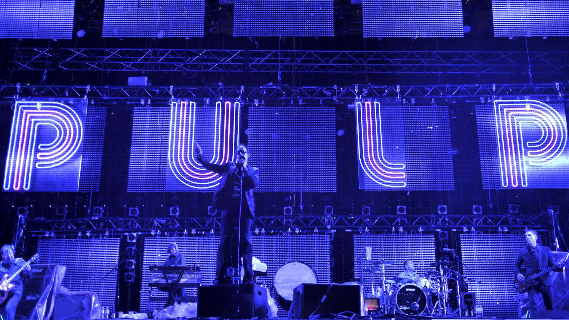 Die Musiker der Band Pulp auf der Bühne vor einer blau angestrahlten Wand. In Leuchtbuchstaben ist "PULP" daran geschrieben. 