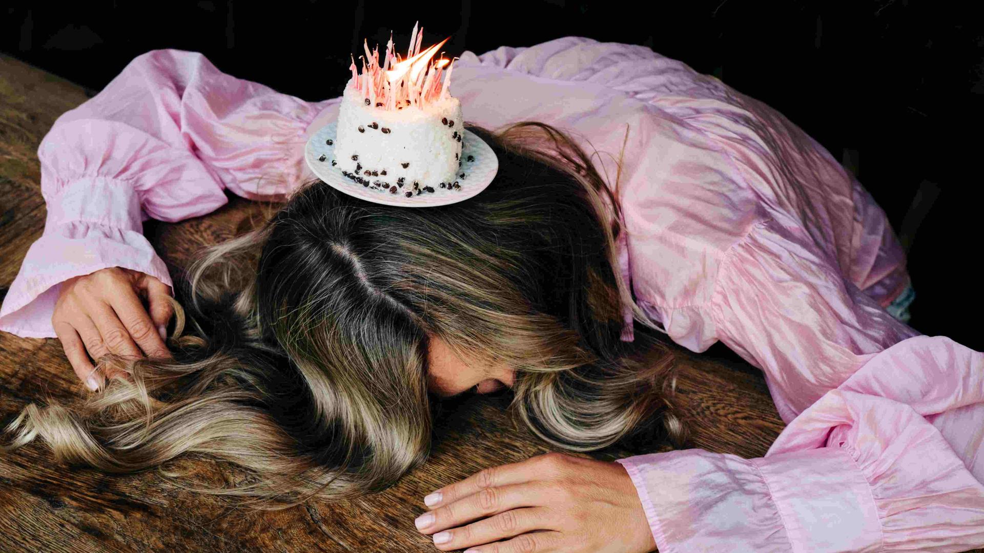 Eine Frau mit pinkfarbener Bluse liegt mit dem Kopf auf einem Holztisch, auf der Hinterseite ihres Kopfs steht ein kleiner ausbalancierter Kuchen mit angezündeten Kerzen.