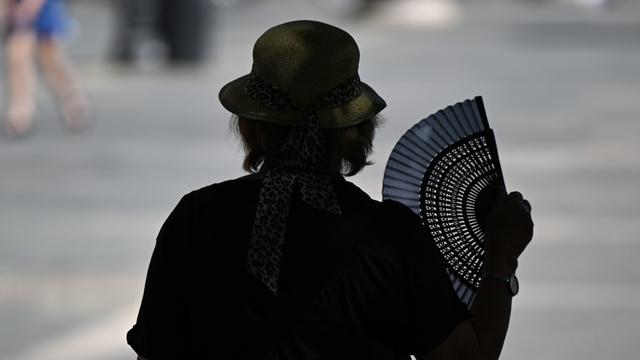Eine ältere Frau ist von hinten im Gegenlicht zu sehen und fächert sich mit einem aufgeklappten Fächer auf dem Frankfurter Opernplatz im Schatten eines Baumes Luft zu.
