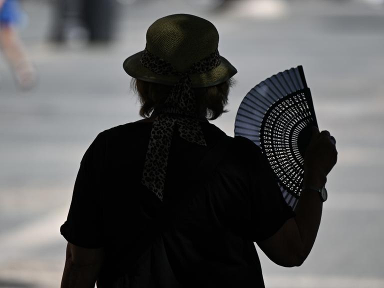 Eine ältere Frau ist von hinten im Gegenlicht zu sehen und fächert sich mit einem aufgeklappten Fächer auf dem Frankfurter Opernplatz im Schatten eines Baumes Luft zu.