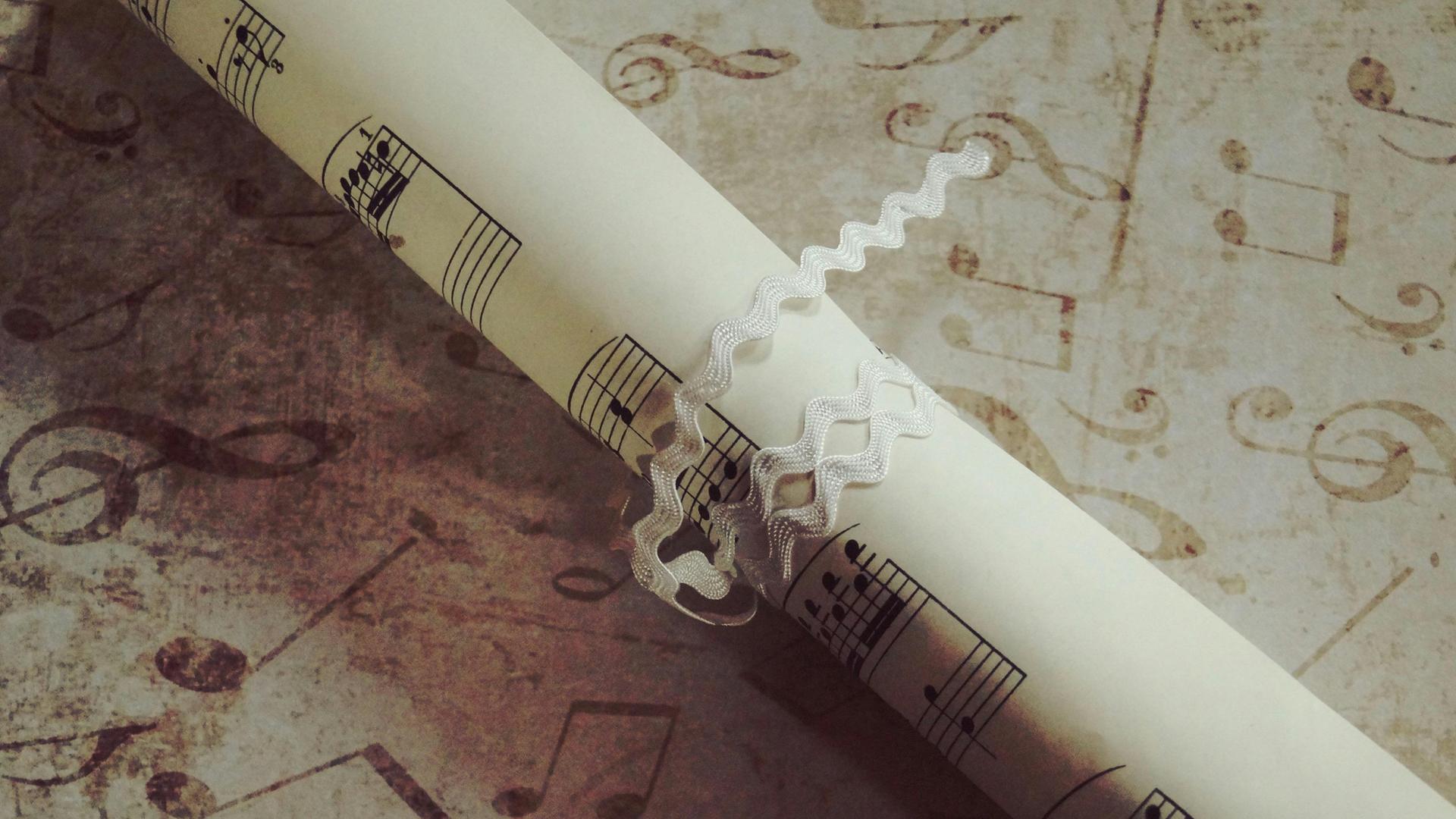 Blick auf Papier, das mit musikalischen Symbolen geschmückt ist, auf dem ein zusammengerolltes Notenblatt mit einem weißen Band liegt.
