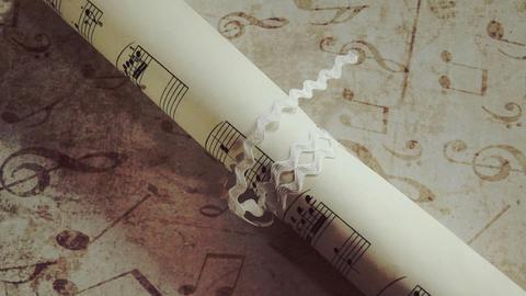 Blick auf Papier, das mit musikalischen Symbolen geschmückt ist, auf dem ein zusammengerolltes Notenblatt mit einem weißen Band liegt.