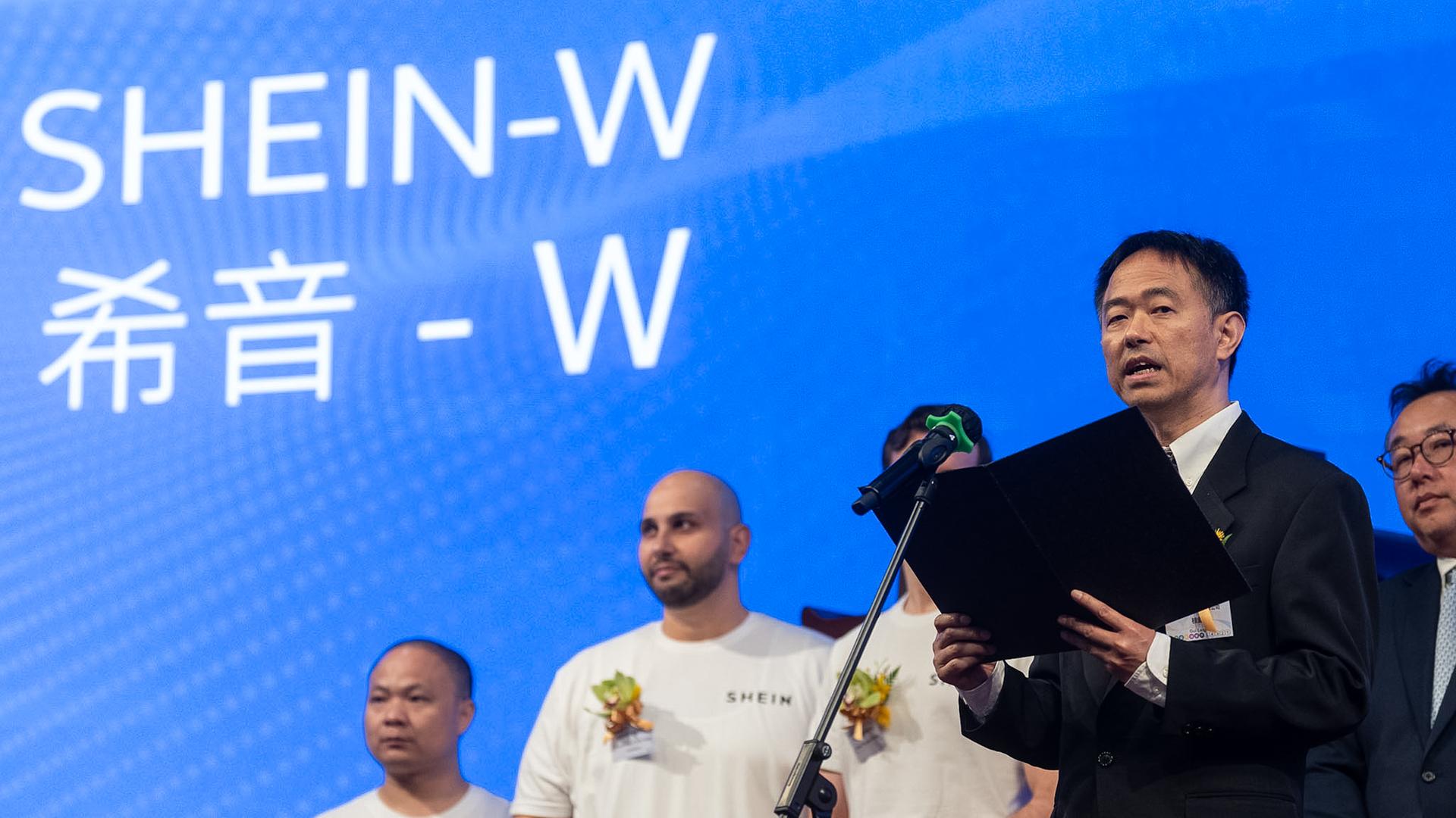Leigh Gui, Finanzvorstand von Shein, spricht während der Zeremonie des Börsengangs. Er steht vor einem Mikrofon und hält eine Mappe in der Hand. Hinter ihm stehen Mitarbeiter. Leigh Gui, Finanzvorstand von Shein, spricht während der Zeremonie des Börsengangs. Er steht vor einem Mikrofon und hält eine Mappe in der Hand. Hinter ihm stehen Mitarbeiter.