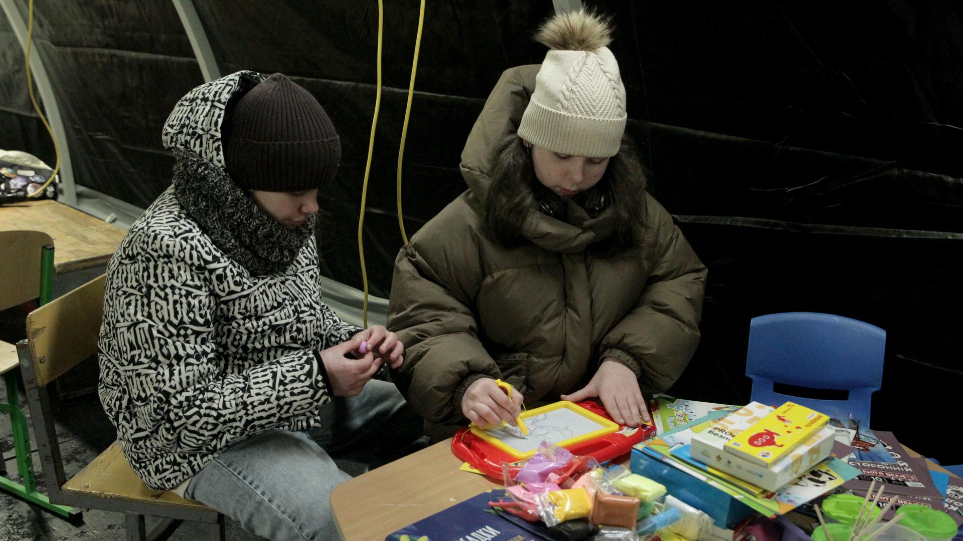Zwei Kinder mit dicken Jacken und mit Mütze sitzen an einem Tisch mit Spielzeug Zwei Kinder mit dicken Jacken und mit Mütze sitzen an einem Tisch mit Spielzeug