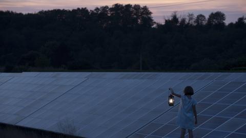 Ein Mädchen steht vor einer Ansammlung von Solarpanelen in der Landschaft und hält eine alte, leuchtende Laterne in der Hand.