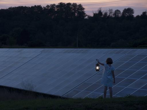 Ein Mädchen steht vor einer Ansammlung von Solarpanelen in der Landschaft und hält eine alte, leuchtende Laterne in der Hand.