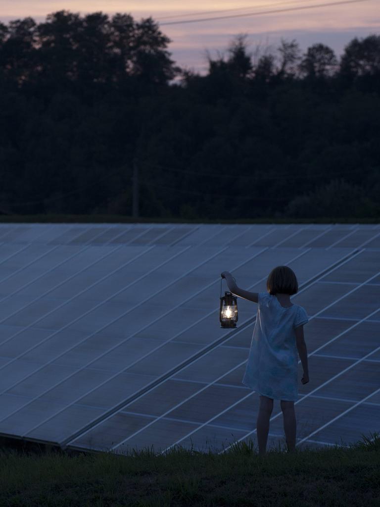 Ein Mädchen steht vor einer Ansammlung von Solarpanelen in der Landschaft und hält eine alte, leuchtende Laterne in der Hand.