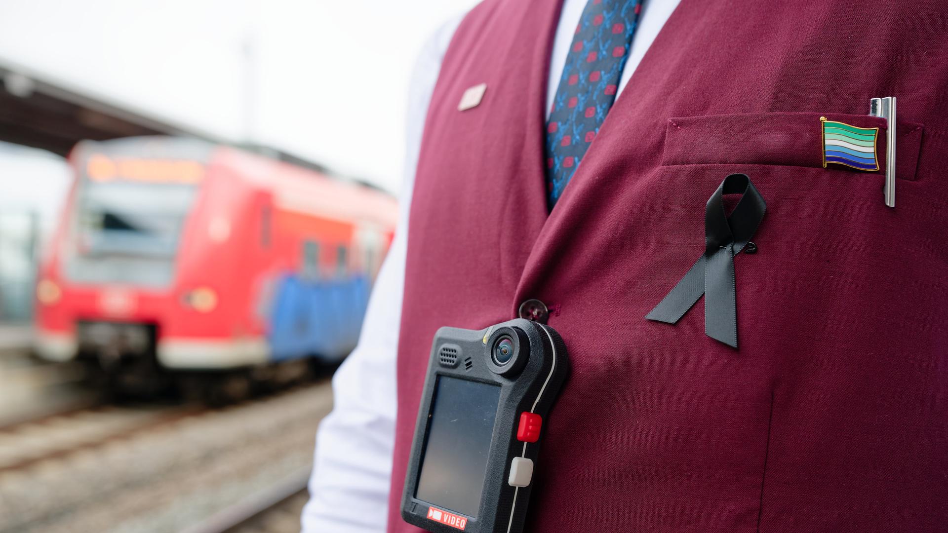 Man sieht den Oberkörper eines Bahn-Mitarbeiters, der eine dunkelrote Weste trägt. Über der Weste hängt an einem schwarzen Band eine sogenannte Bodycam. Im Hintergrund sieht man einen roten Regionalzug der Deutschen Bahn.