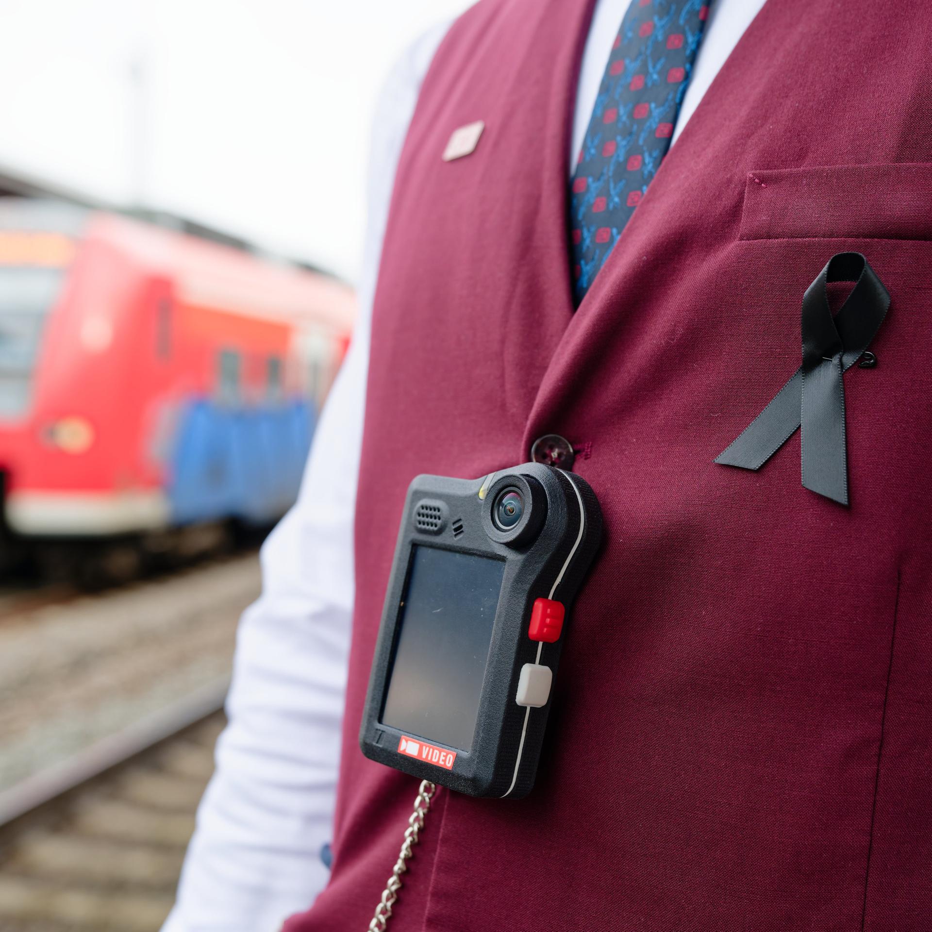 Man sieht den Oberkörper eines Bahn-Mitarbeiters, der eine dunkelrote Weste trägt. Über der Weste hängt an einem schwarzen Band eine sogenannte Bodycam. Im Hintergrund sieht man einen roten Regionalzug der Deutschen Bahn.
