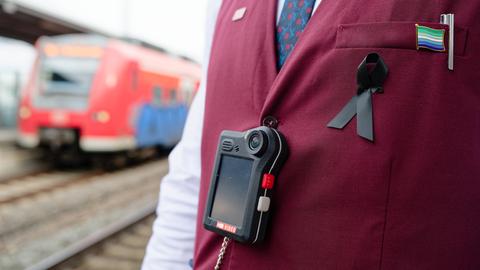 Der Oberkörper eines Bahn-Mitarbeiters, der eine dunkelrote Weste trägt. Über der Weste hängt an einem schwarzen Band eine sogenannte Bodycam. Im Hintergrund sieht man einen roten Regionalzug der Deutschen Bahn.