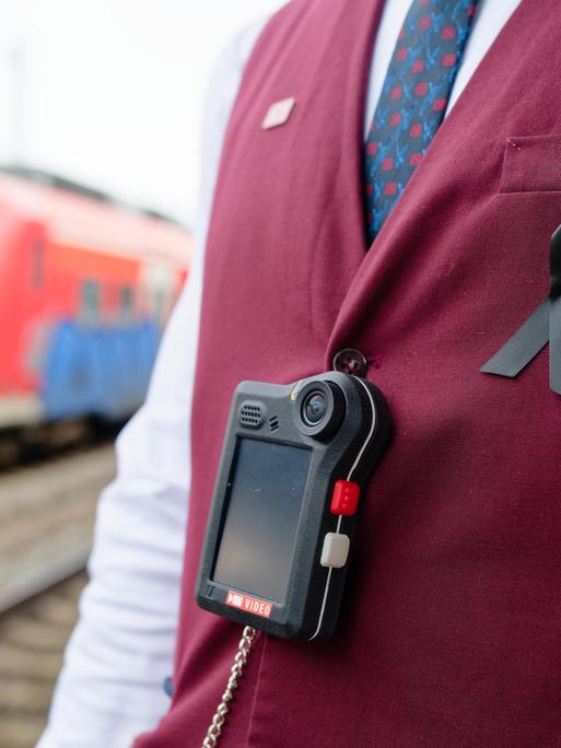 Man sieht den Oberkörper eines Bahn-Mitarbeiters, der eine dunkelrote Weste trägt. Über der Weste hängt an einem schwarzen Band eine sogenannte Bodycam. Im Hintergrund sieht man einen roten Regionalzug der Deutschen Bahn. Man sieht den Oberkörper eines Bahn-Mitarbeiters, der eine dunkelrote Weste trägt. Über der Weste hängt an einem schwarzen Band eine sogenannte Bodycam. Im Hintergrund sieht man einen roten Regionalzug der Deutschen Bahn.