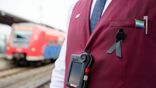 Der Ober-Körper von einem Bahn-Mitarbeiter. Über seiner Weste hängt an einem schwarzen Band eine Body-Cam. Im Hinter-Grund ein roter Regional-Zug der Deutschen Bahn.