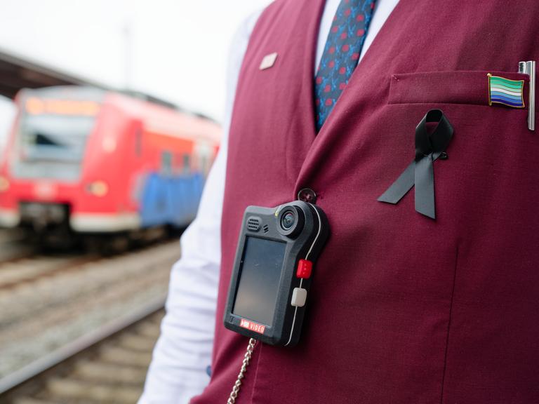 Man sieht den Oberkörper eines Bahn-Mitarbeiters, der eine dunkelrote Weste trägt. Über der Weste hängt an einem schwarzen Band eine sogenannte Bodycam. Im Hintergrund sieht man einen roten Regionalzug der Deutschen Bahn. Man sieht den Oberkörper eines Bahn-Mitarbeiters, der eine dunkelrote Weste trägt. Über der Weste hängt an einem schwarzen Band eine sogenannte Bodycam. Im Hintergrund sieht man einen roten Regionalzug der Deutschen Bahn.