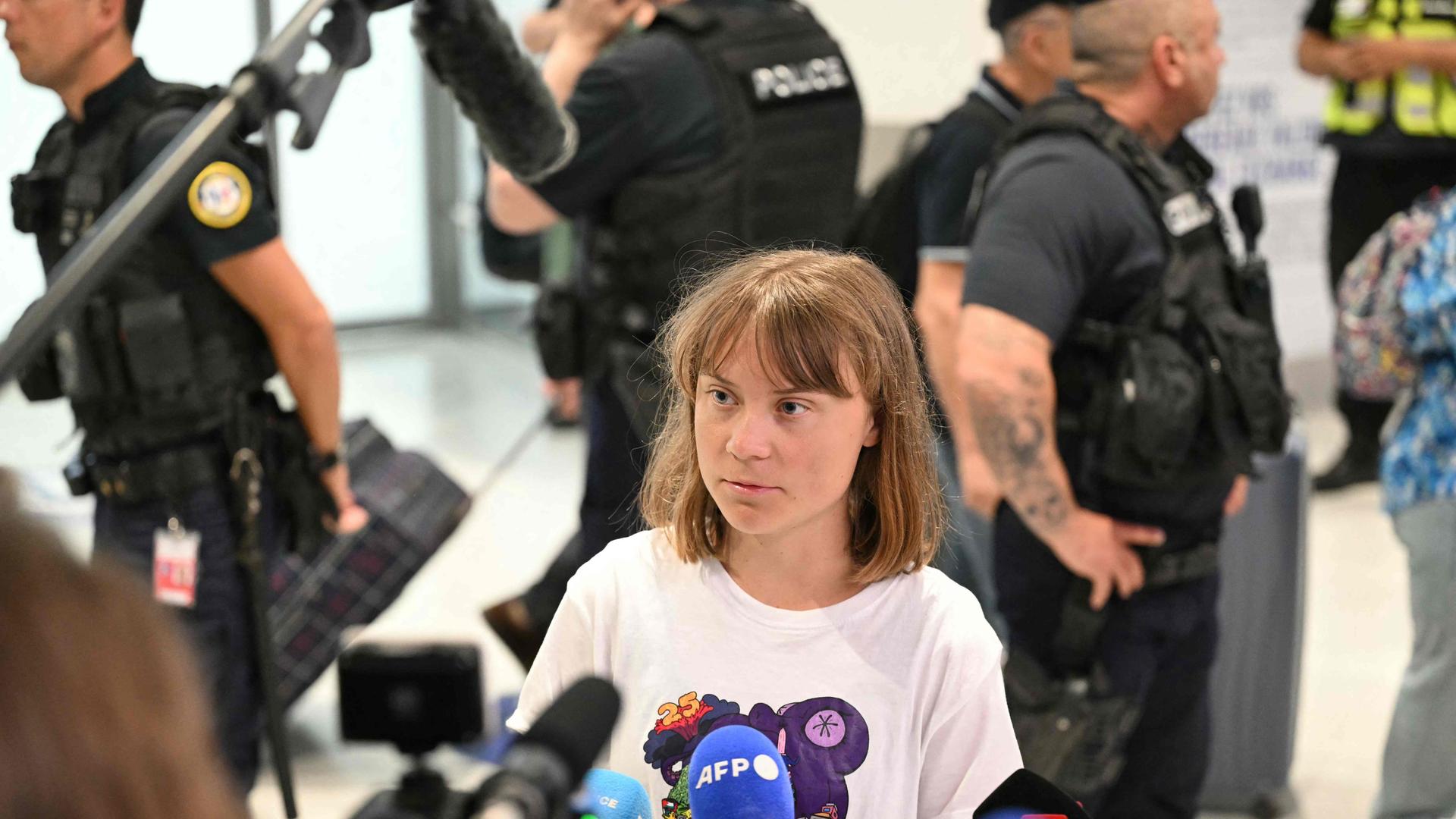 Thunberg steht in einem Flughafengebäude in einem weißen T-Shirt vor mehreren Mikrofonen. Dahinter mehrere Polizisten. 