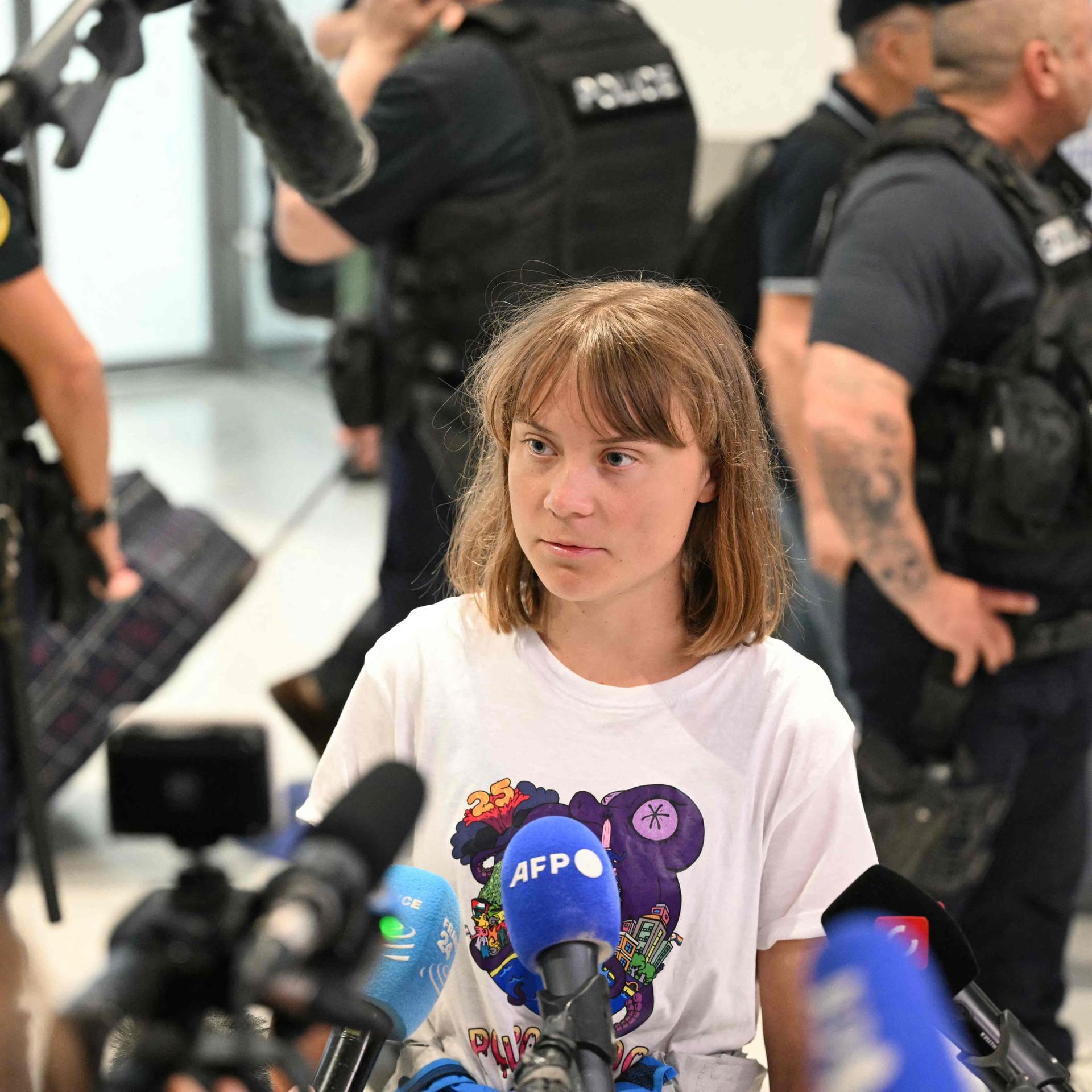 Thunberg steht in einem Flughafengebäude in einem weißen T-Shirt vor mehreren Mikrofonen. Dahinter mehrere Polizisten. Thunberg steht in einem Flughafengebäude in einem weißen T-Shirt vor mehreren Mikrofonen. Dahinter mehrere Polizisten.