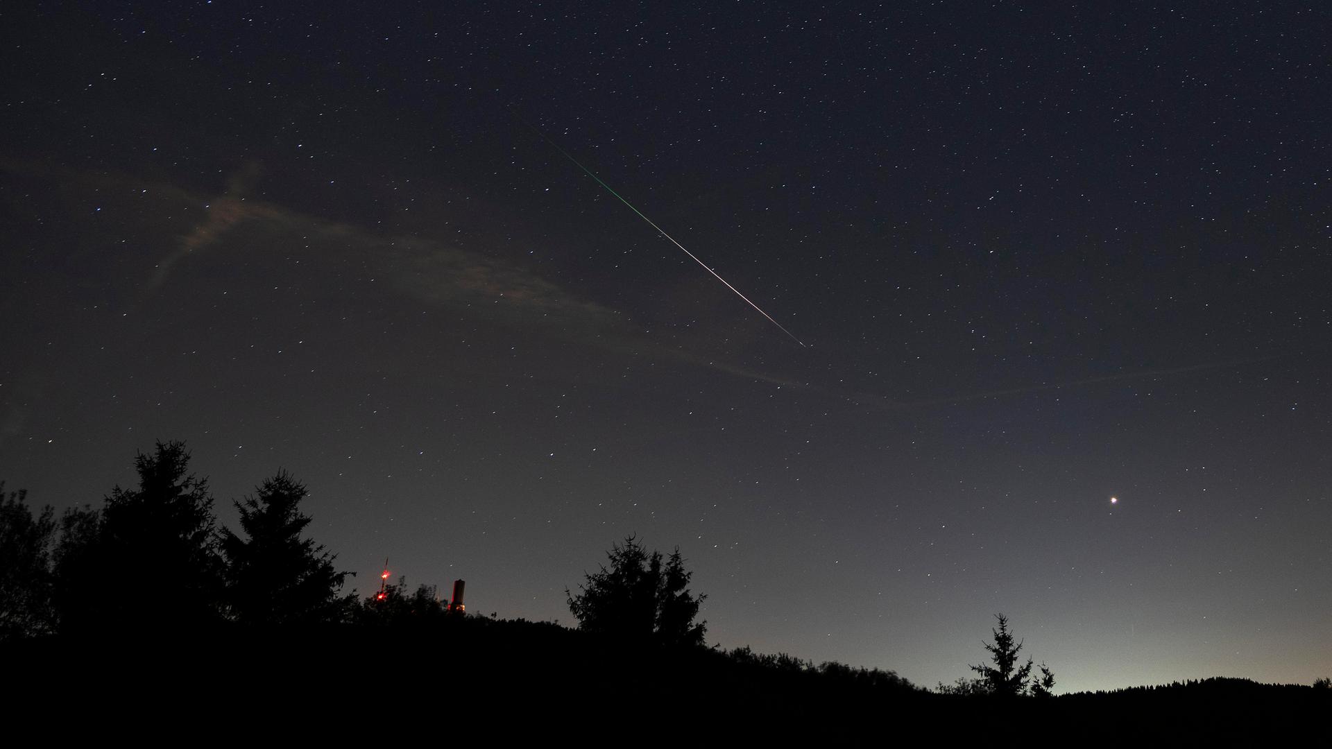 Über einem Wald ist am Himmel der helle Streifen einer Stern-Schnuppe zu erkennen. Über einem Wald ist am Himmel der helle Streifen einer Stern-Schnuppe zu erkennen.