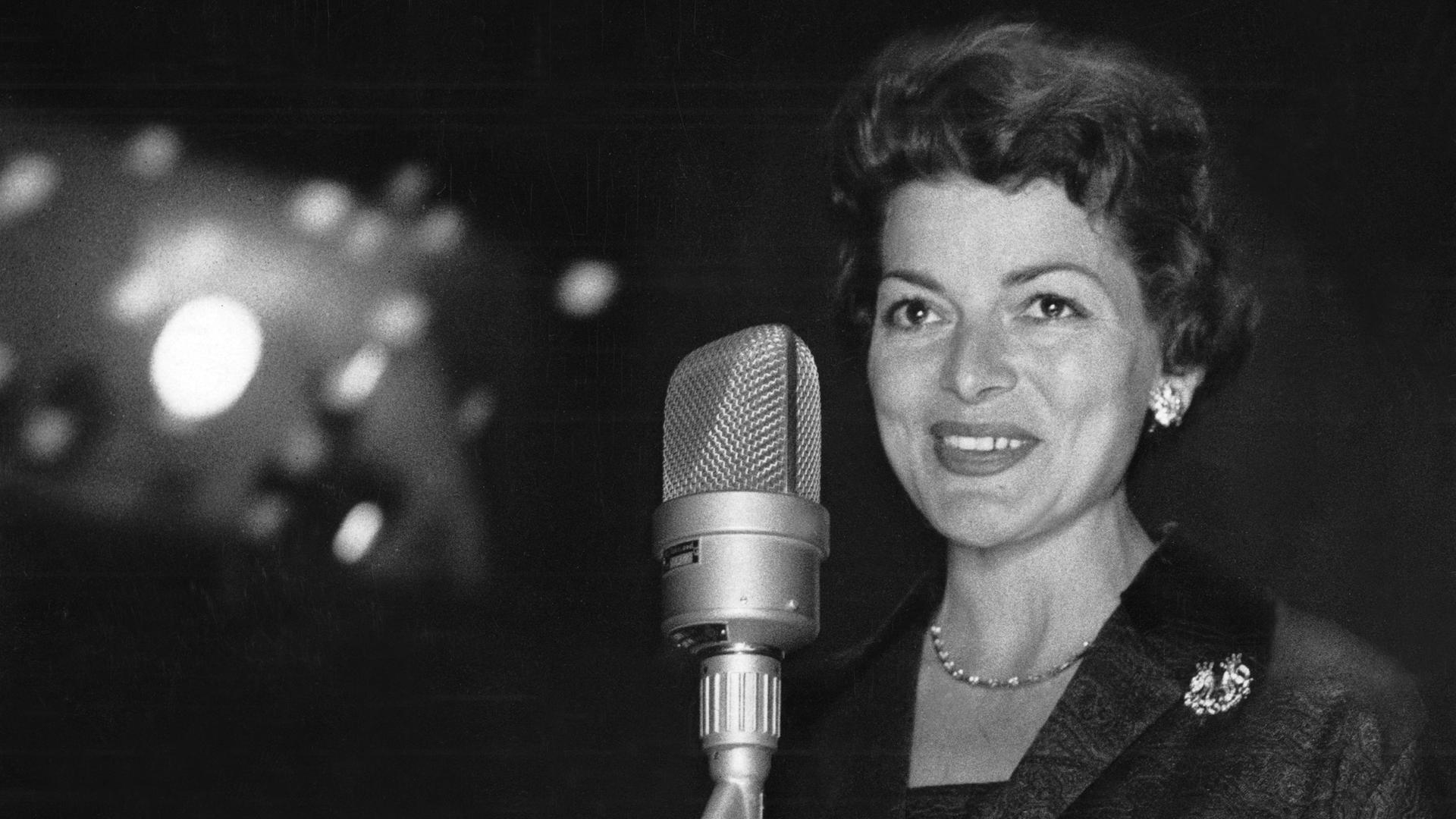 Lys Assia posiert vor einem dunklen Hintergrund hinter einem Mikrofon für ein Schwarz-Weiß-Pressefoto.