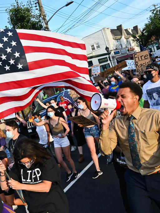 Demonstranten während einer Black-Lives-Matter-Demonstration tragen eine amerikanische Flagge und schreien wütend in ein Megafon. Demonstranten während einer Black-Lives-Matter-Demonstration tragen eine amerikanische Flagge und schreien wütend in ein Megafon.