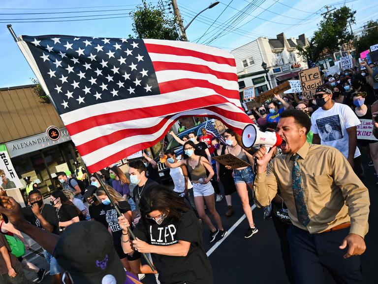 Demonstranten während einer Black-Lives-Matter-Demonstration tragen eine amerikanische Flagge und schreien wütend in ein Megafon. Demonstranten während einer Black-Lives-Matter-Demonstration tragen eine amerikanische Flagge und schreien wütend in ein Megafon.