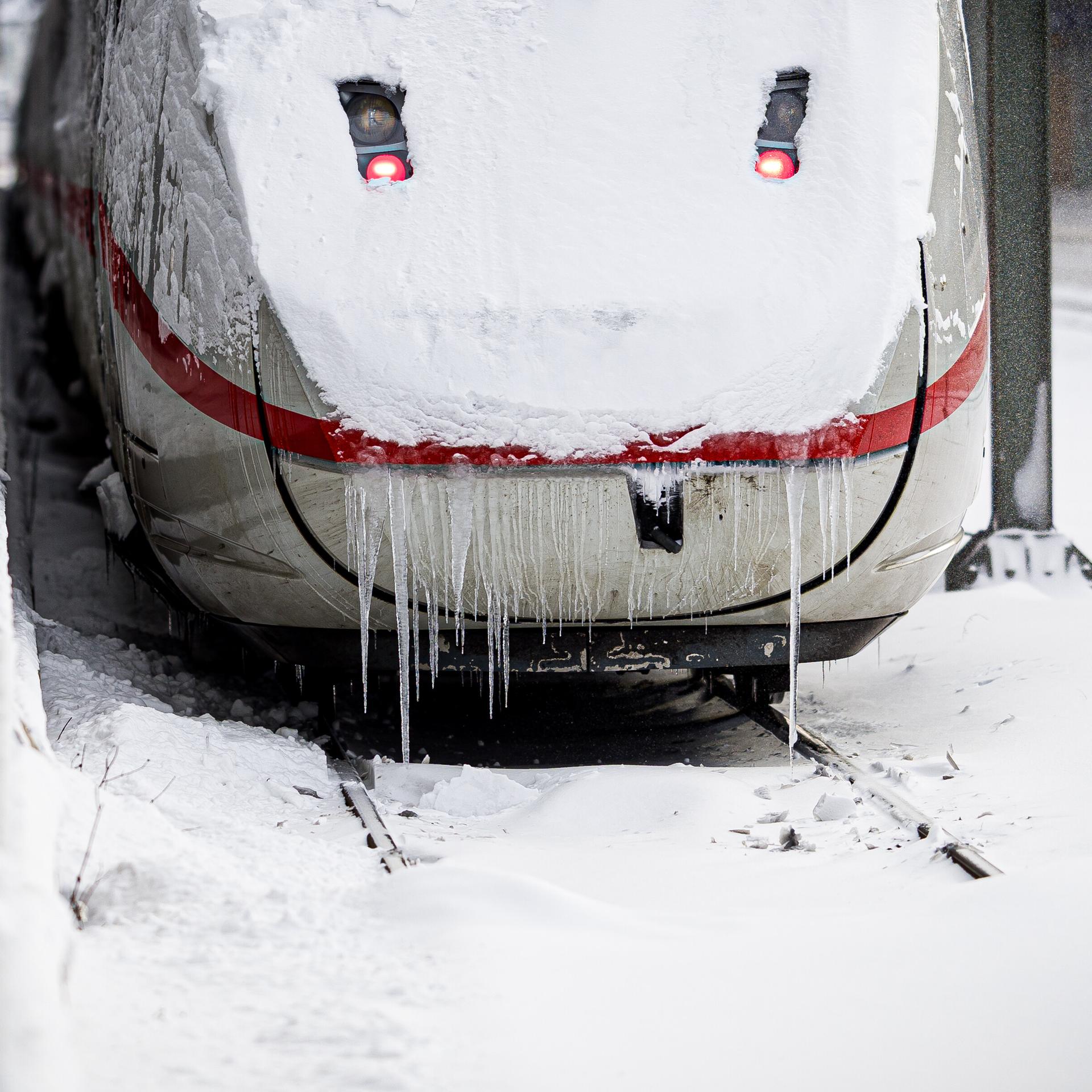 Rückansicht eines ICE-Zuges, dessen Scheibe verschneit ist. Lange Eiszapfen reichen bis auf die Schienen.