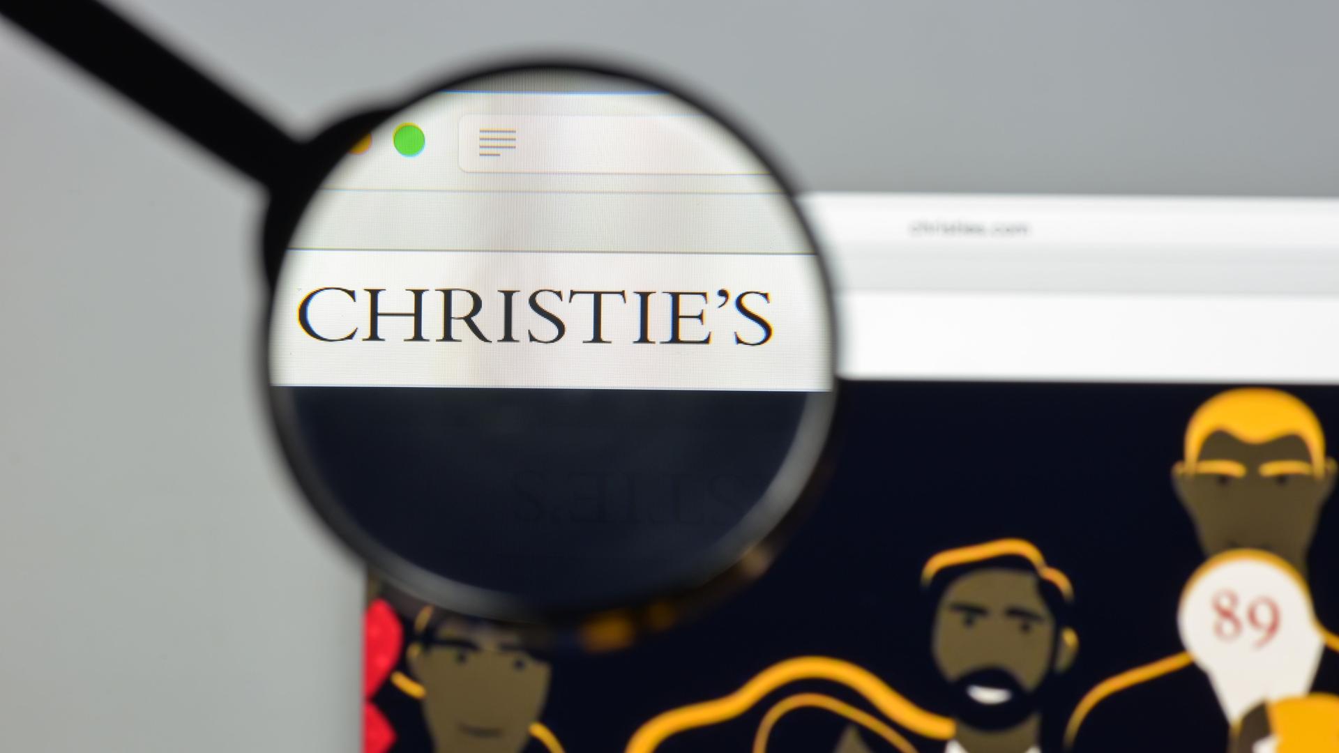 Die Website des britischen Auktionshaus Christie's ist auf einem Laptop geöffnet. Eine Hand hält eine Lupe über das Logo, das scharf dargestellt wird, der Rest erscheint unscharf.