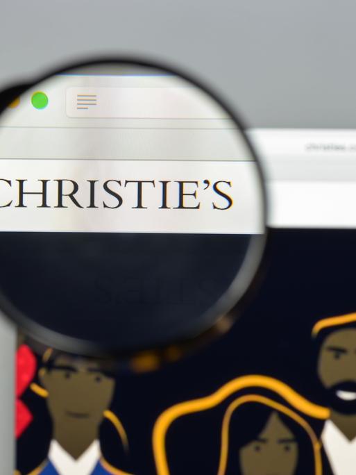 Die Website des britischen Auktionshaus Christie's ist auf einem Laptop geöffnet. Eine Hand hält eine Lupe über das Logo, das scharf dargestellt wird, der Rest erscheint unscharf.