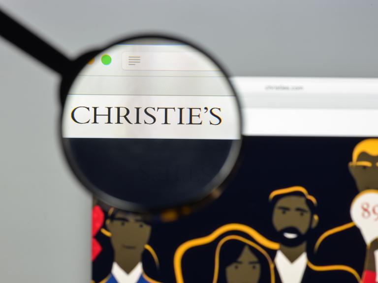 Die Website des britischen Auktionshaus Christie's ist auf einem Laptop geöffnet. Eine Hand hält eine Lupe über das Logo, das scharf dargestellt wird, der Rest erscheint unscharf.