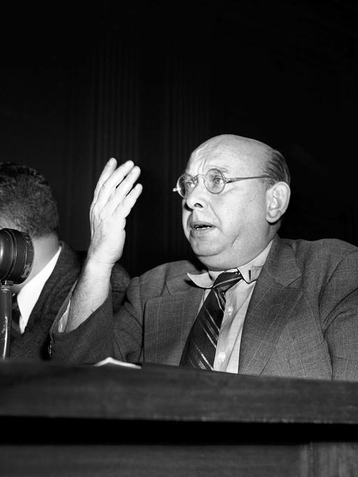 Das Schwarz-Weiß-Foto zeigt einen Mann in Tweet-Anzug und mit Nickelbrille, der mit der rechten Hand gestikuliert