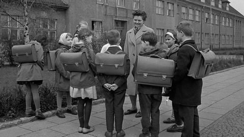 Historisches Foto: Eine Lehrerin und Schüler stehen mit Tornister, Schulranzen, Schultasche vor einer Schule.