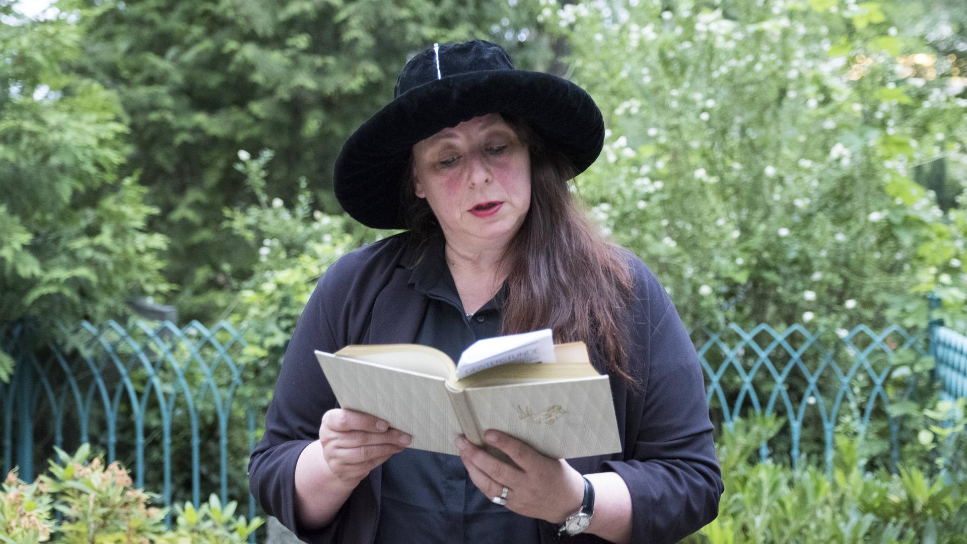 Annett Gröschner mit schwarzem Hut und schwarzem Oberteil liest vor grüner Kulisse aus einem Buch.