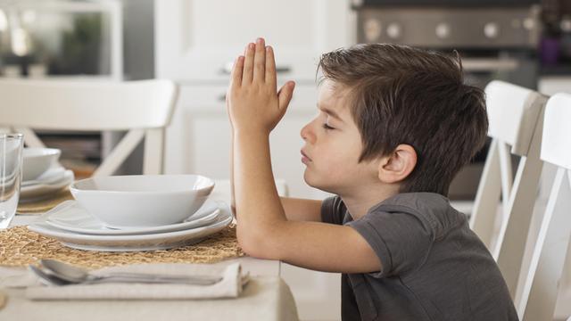 Ein kleiner Junge sitzt am Esstisch vor einem leeren Teller. Er betet.