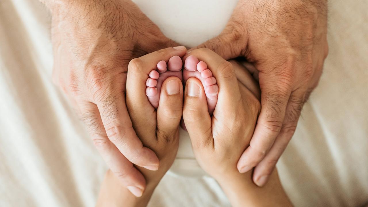 Die Hände eines älteren Mannes umfassen die Hand einer Frau, die wiederum die Füße eines Babys umfasst.