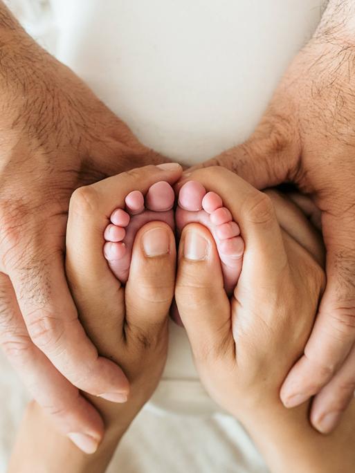 Die Hände eines älteren Mannes umfassen die Hand einer Frau, die wiederum die Füße eines Babys umfasst.