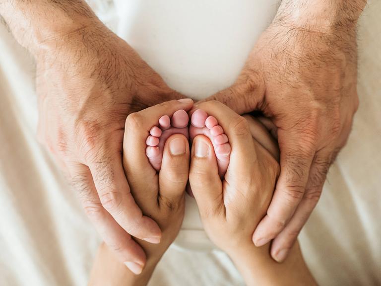 Die Hände eines älteren Mannes umfassen die Hand einer Frau, die wiederum die Füße eines Babys umfasst.