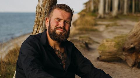 Jan Gorkow alias Monchi, Sänger von „Feine Sahne Fischfilet“, lehnt an einem Strand an einem abgestorbenen Baumstamm und blickt in die Kamera.