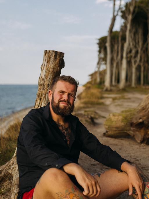 Jan Gorkow alias Monchi, Sänger von „Feine Sahne Fischfilet“, lehnt an einem Strand an einem abgestorbenen Baumstamm und blickt in die Kamera.