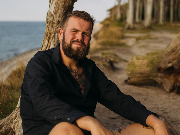 Jan Gorkow alias Monchi, Sänger von „Feine Sahne Fischfilet“, lehnt an einem Strand an einem abgestorbenen Baumstamm und blickt in die Kamera.