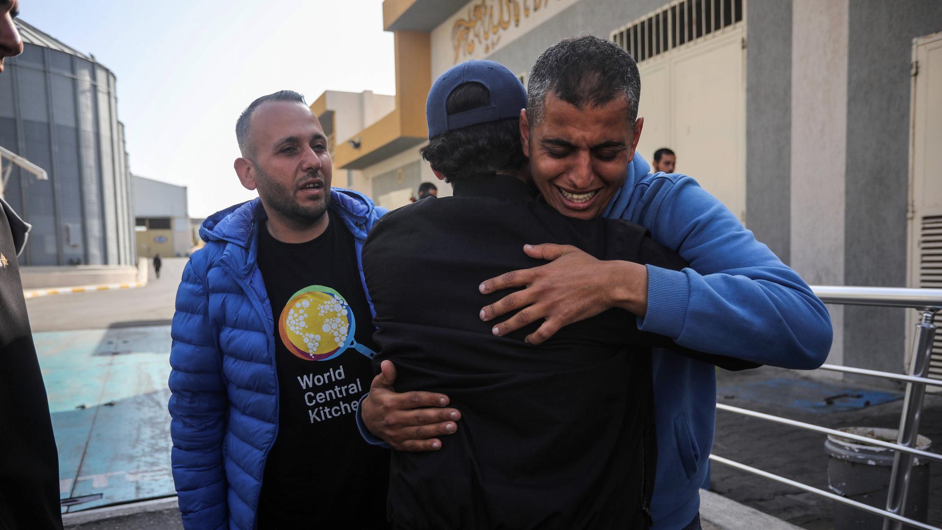 Drei Männer trauern um in Gaza die getöteten Helfer der Organisation World Central Kitchen. Zwei von iohnen umarmen sich. 